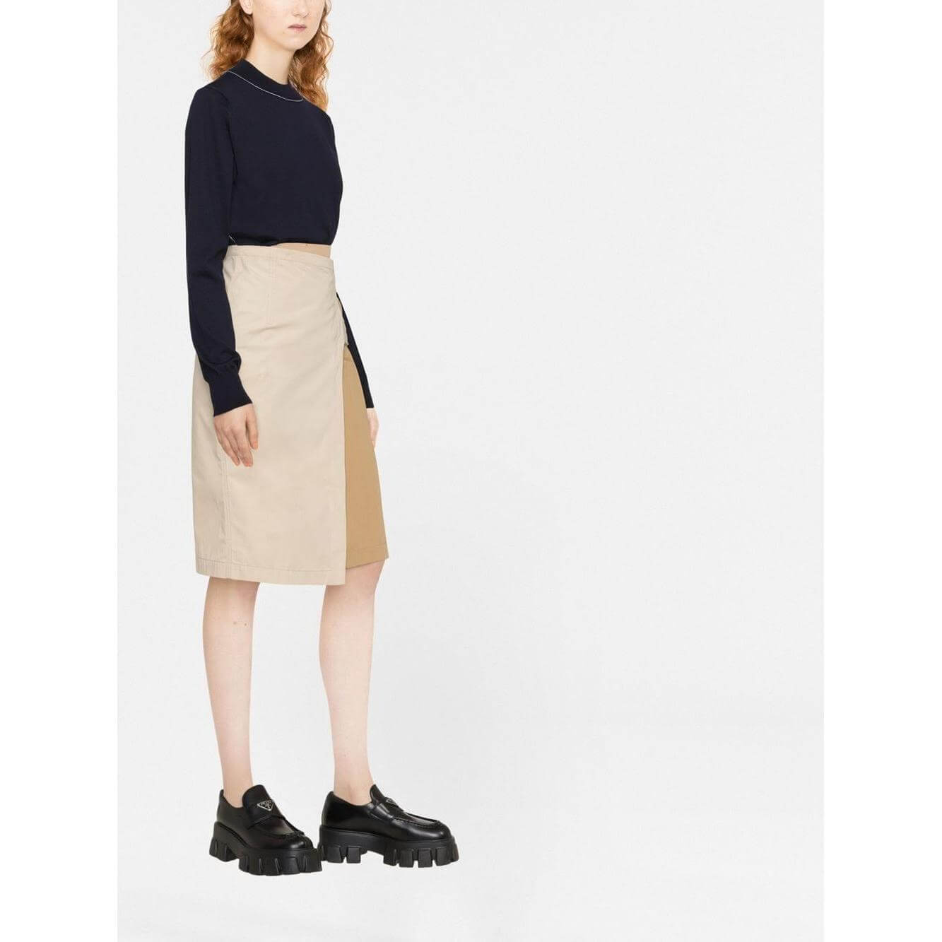 Woman wearing navy Maison Margiela sweater and beige skirt standing on white background.