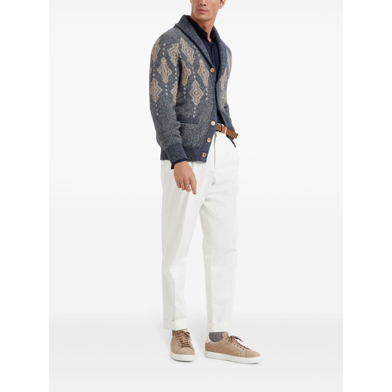 Man wearing a stylish button-up cardigan with pattern, navy shirt, white trousers, and tan sneakers, standing against a plain background.