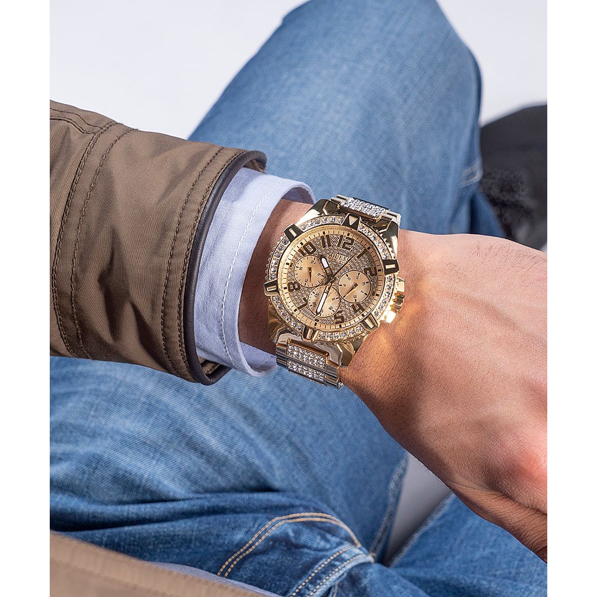 Alt Tag: Person wearing a luxury GUESS Mod. FRONTIER watch with gold detailing, styled with casual blue jeans and a brown jacket.