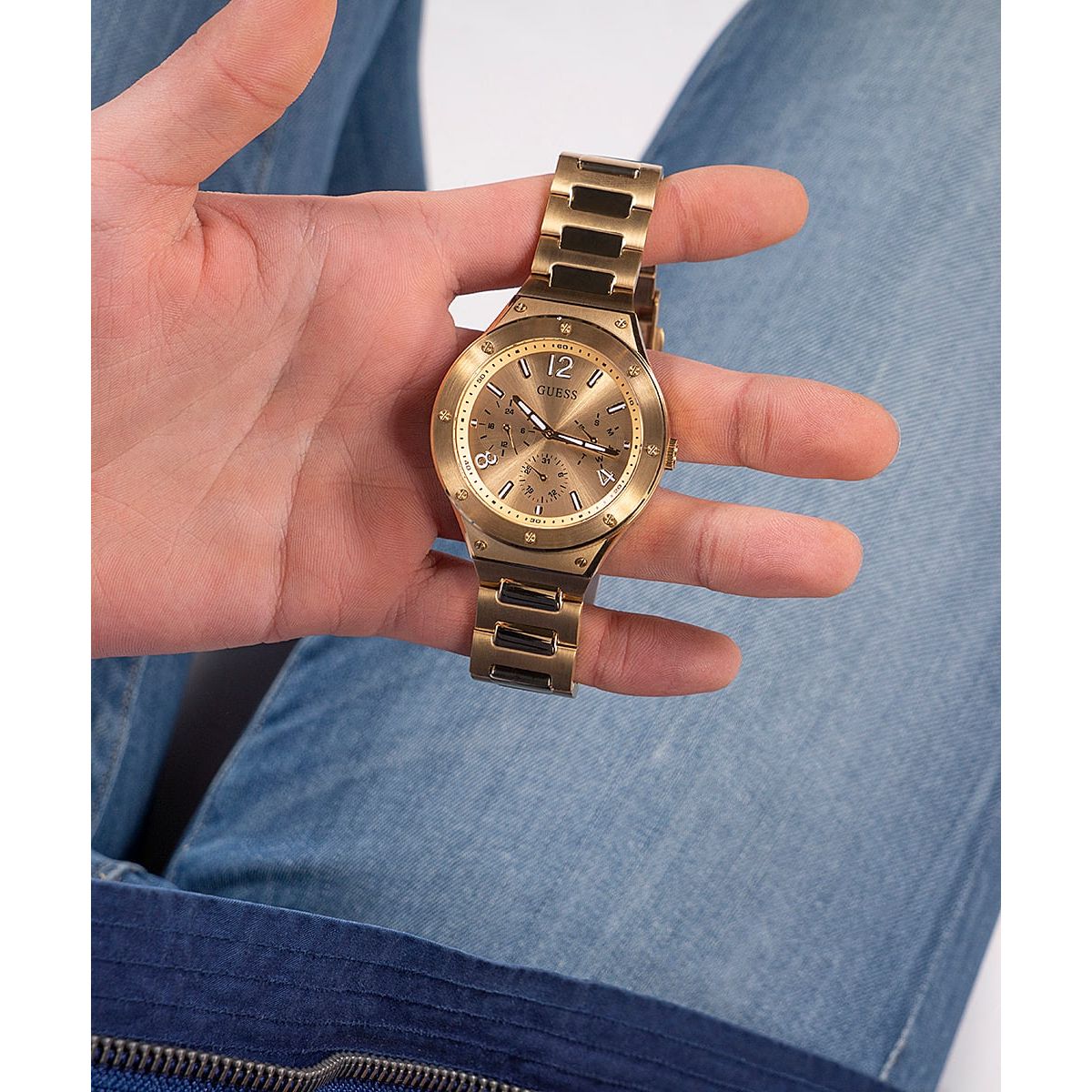 Person holding gold GUESS watch with quartz analog movement, featuring mineral dial and 5 ATM water resistance.